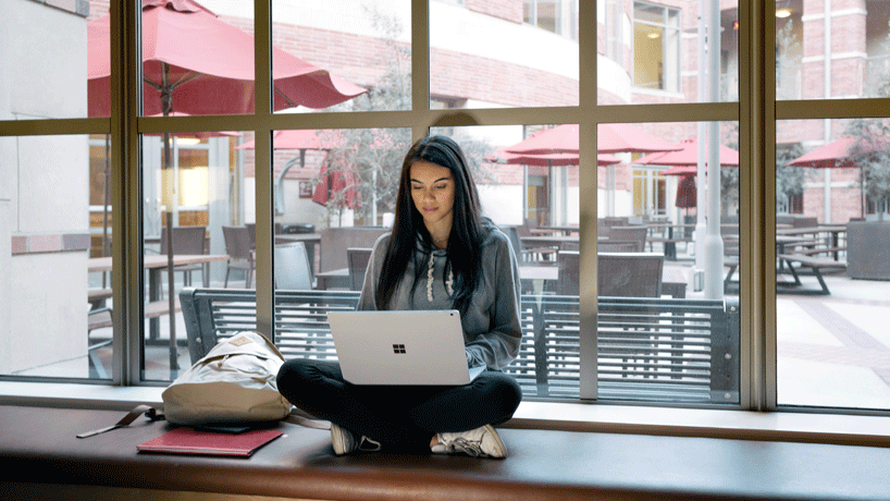 Surface ノート PC で認定Microsoft 365 開発者になるために研究を行う、何気なく脚を組んで座っている女性
